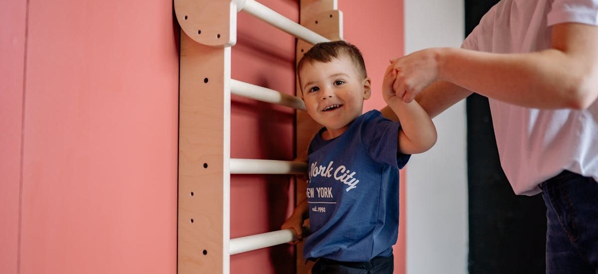 A smiling boy climbs a wooden ladder with a woman's assistance, embodying teamwork and joy indoors.