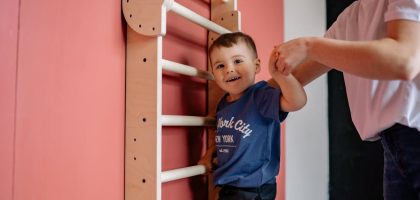 A smiling boy climbs a wooden ladder with a woman's assistance, embodying teamwork and joy indoors.