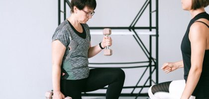 Elderly woman lifting dumbbells with guidance from a trainer indoors.
