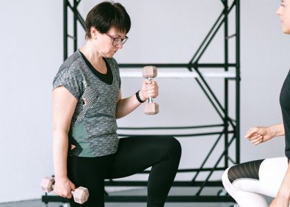 Elderly woman lifting dumbbells with guidance from a trainer indoors.